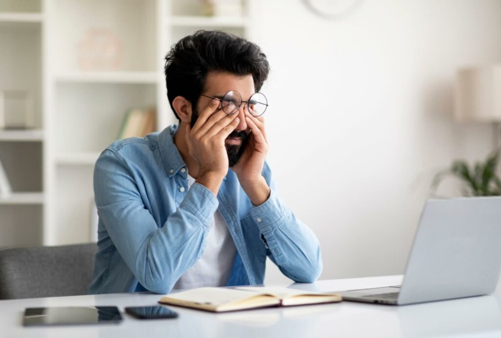 Young adult man rubbing his eyes while at computer.