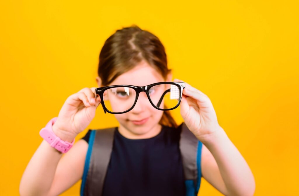 Against a yellow background, a school-age child holds up a pair of eyeglasses that help them read in class.