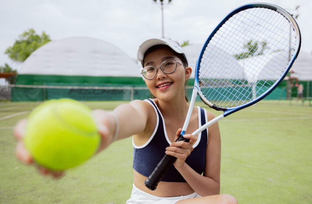 A teenager wearing glasses to correct myopia holds a tennis ball and racket while practicing the sport.