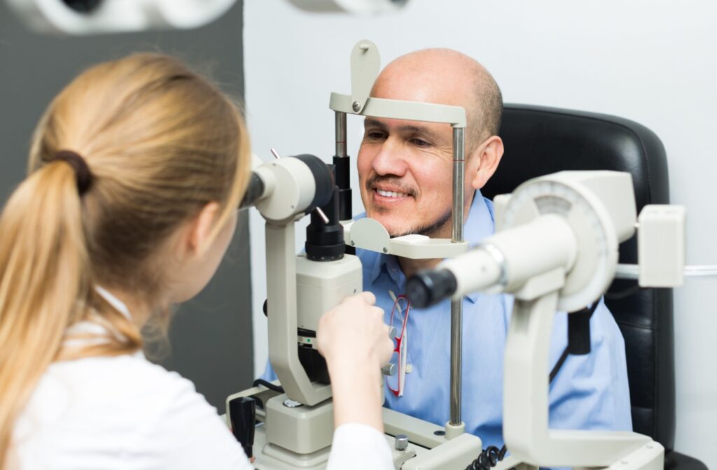 An optometrist performing an eye exam on a smiling adult male patient using a slit lamp machine in an exam room.