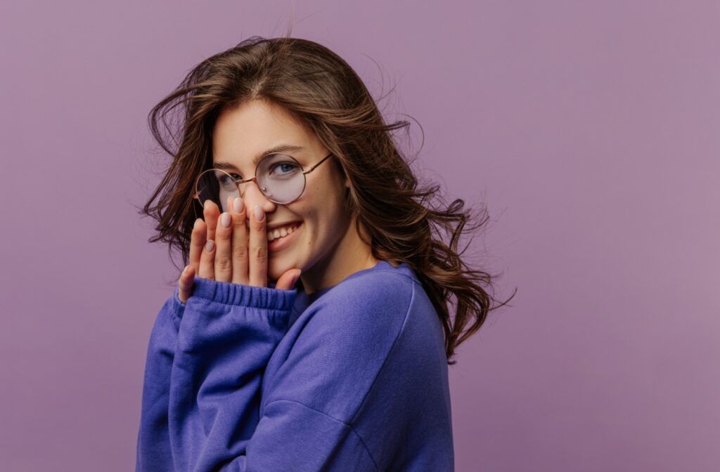 Smiling woman in round glasses and blue sweater poses playfully against a purple background.