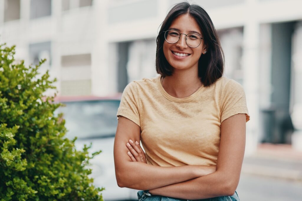 Smiling young woman wearing prescription eyeglasses standing with arms crossed in bright indoor setting.
