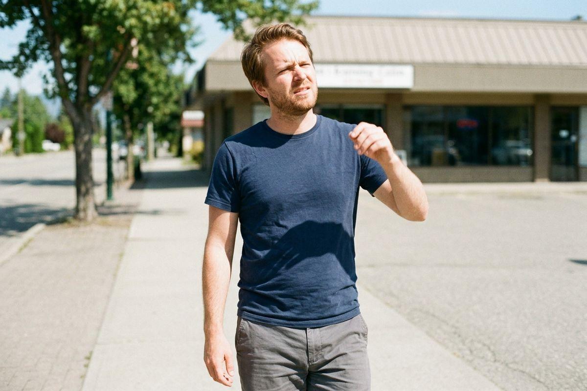 Adult in navy t-shirt squinting while walking on a sunny sidewalk in front of storefronts.